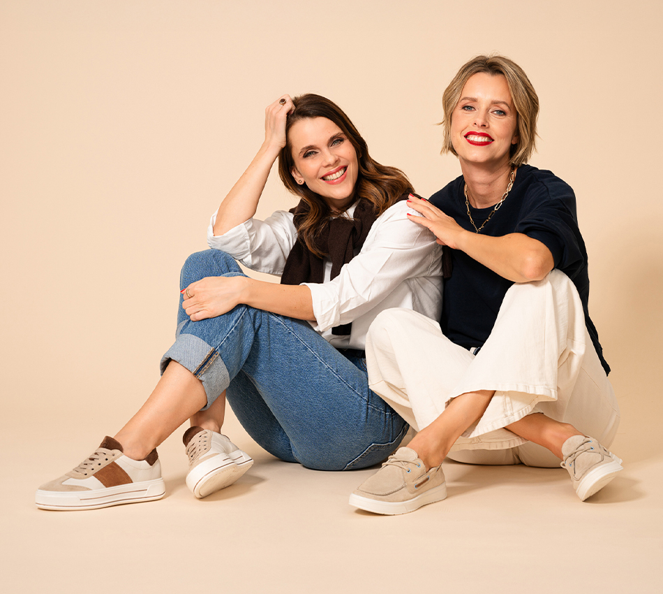 Zwei Frauen sitzen lächelnd auf dem Boden und präsentieren helle ara Sneaker und Slipper vor einem neutralen beigen Hintergrund.